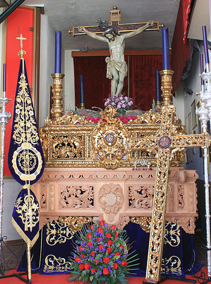 La imagen del Sant&iacute;simo Cristo Crucificado de Padul expuesta en su paso procesional dorado y tallado en madera, adornado con arreglos florales en tonos morados y rojos, flanqueado por el estandarte bordado y la cruz de gu&iacute;a dorada durante la ma&ntilde;ana del Viernes Santo.
